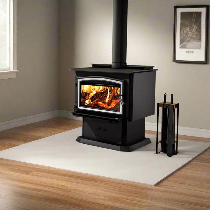 A black wood-burning stove with a visible fire inside, set on a white rug in a room with wood floors and beige walls. A matching black fireplace tool stand is beside it.