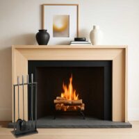 A light beige wood mantel and fireplace surround with a black firebox, shown with a burning fire and decorative items on the shelf above.