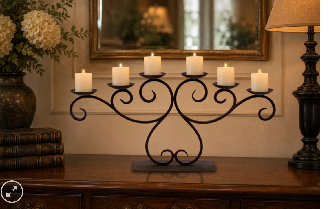 A black wrought-iron candelabra with five candle holders, shown lit with five white pillar candles on a wooden console table in a warmly lit room.