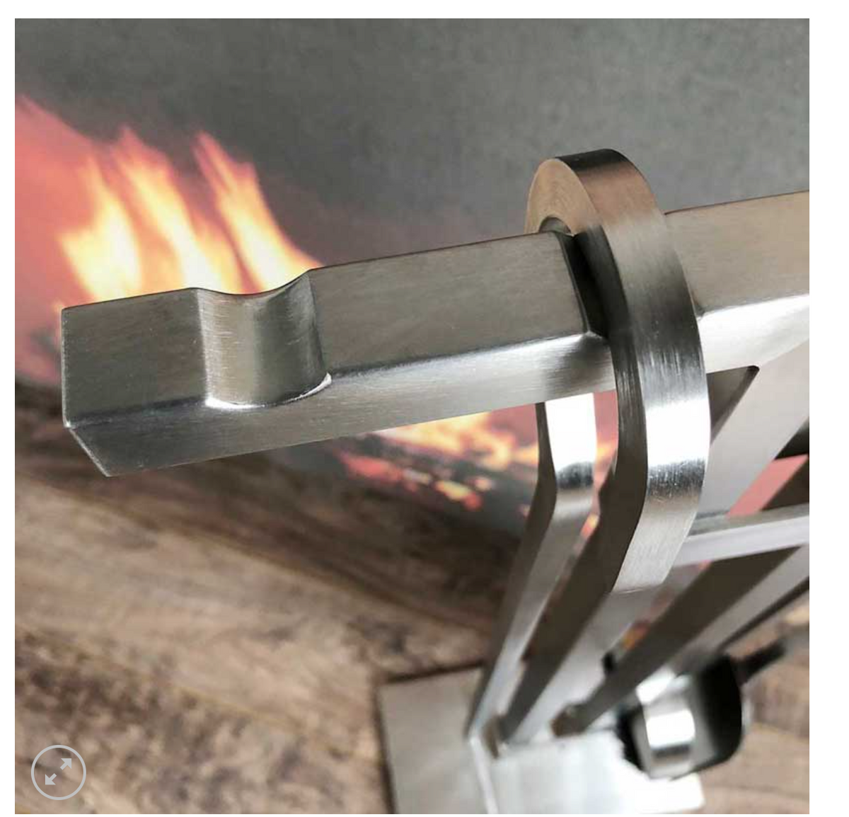 A close-up of a brushed stainless steel fireplace poker resting on a metal stand, with blurred flames and a wood-textured surface in the background.