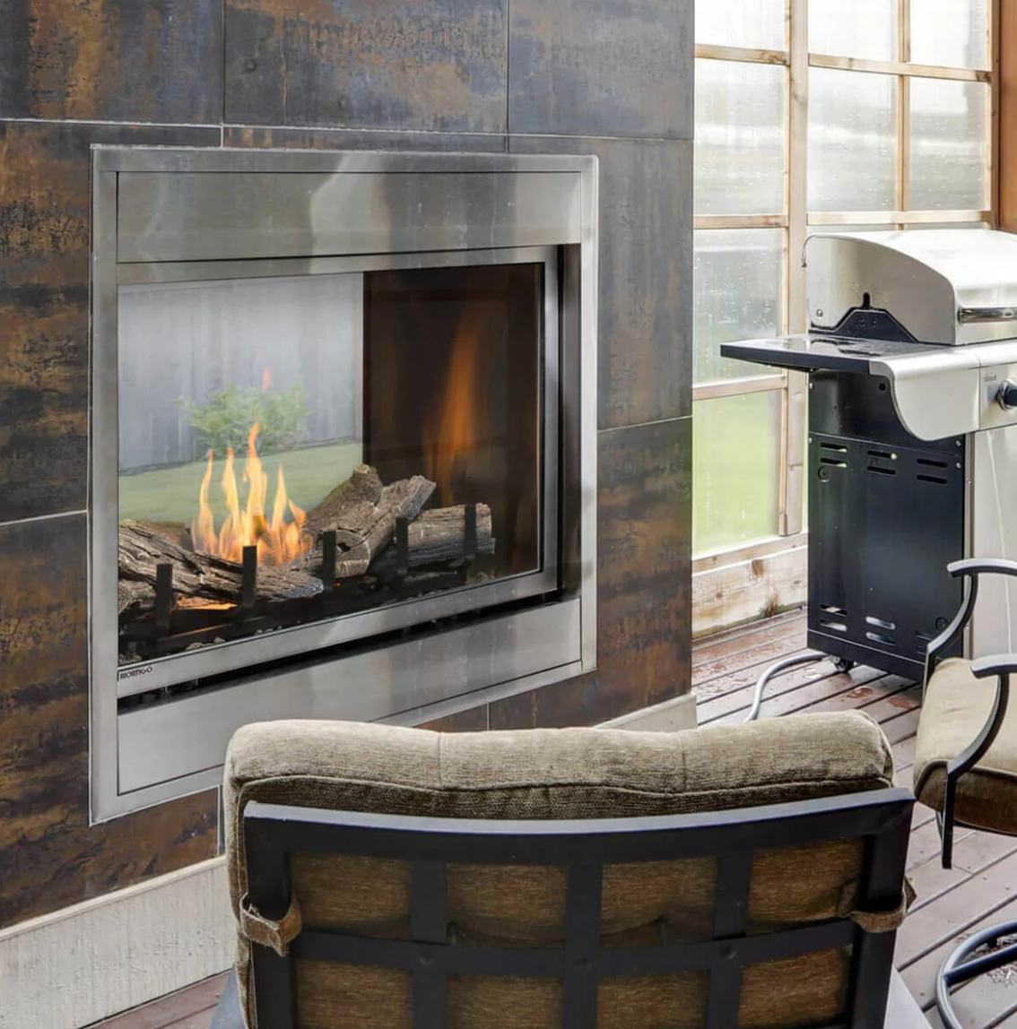 fireplace in outdoor setting with a grill and chairs in the background. Fireplace is see through built into a dtone look wall