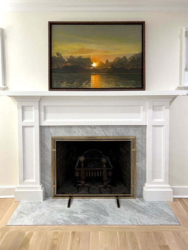 A framed landscape painting of a sunset over water hangs on a white wall above a white fireplace mantel with a gray marble hearth and gold fire screen.