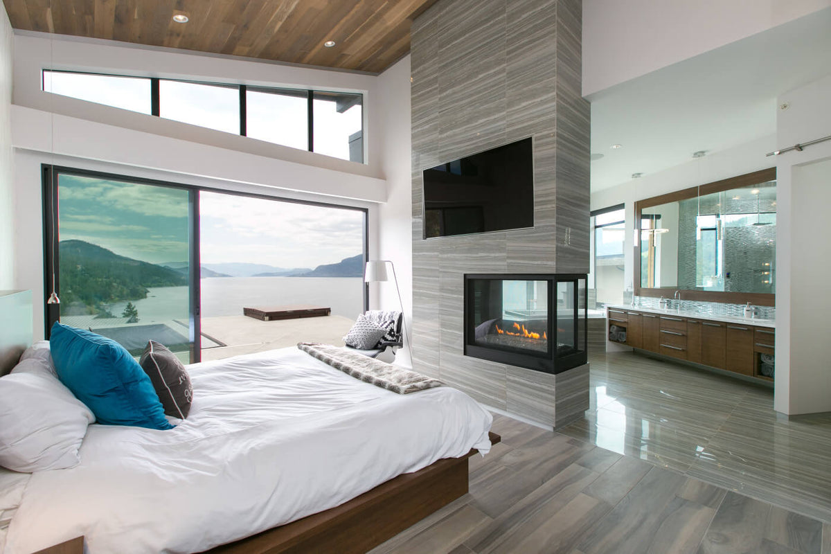 A modern bedroom interior with a large bed dressed in white bedding and blue accent pillows, set in a high-ceilinged room with wood flooring, a stone fireplace, and a glass wall overlooking water and mountains. A bathroom is visible in the background.