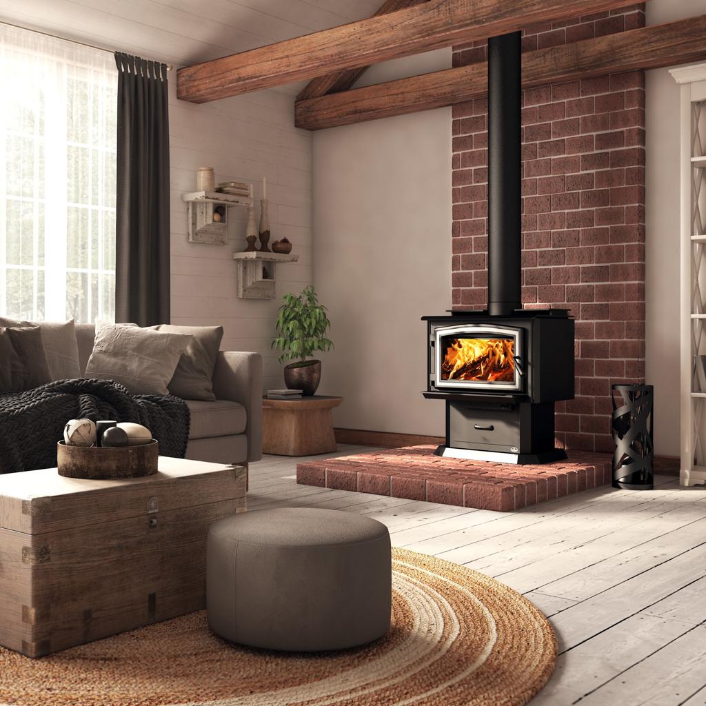 A black wood-burning stove with a visible fire, set against a red brick wall in a rustic living room with a gray sofa, wooden ceiling beams, and a round ottoman.