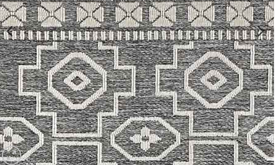 A close-up of a woven rug or textile with a geometric pattern in gray and off-white, featuring large diamond and cross motifs with a decorative border at the top.
