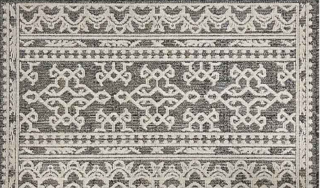 An area rug with a distressed geometric pattern in charcoal gray and off-white, featuring ornate borders and central medallions.