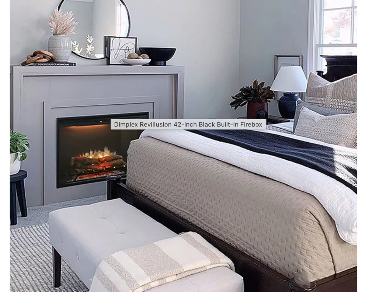 A built-in fireplace installed in a light gray fireplace mantel, shown in a styled bedroom setting with a bed, bench, lamp, and window. A text overlay reads 'Dimplex Revillusion 42-inch Black Built-In Firebox.