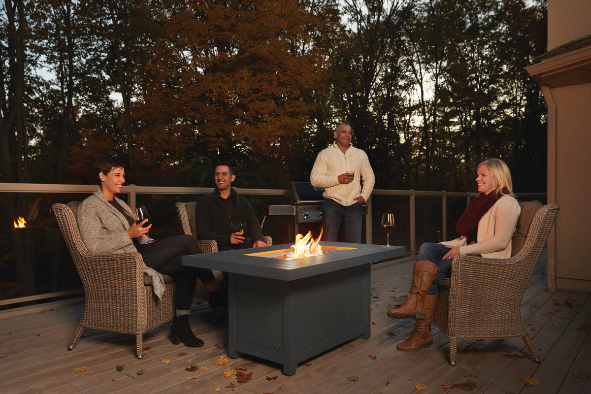 Four people gathered on a wooden deck at dusk around a square gray fire pit table, seated in wicker-style armchairs. Trees with autumn foliage fill the background, and the fire pit flame glows in the center of the table.
