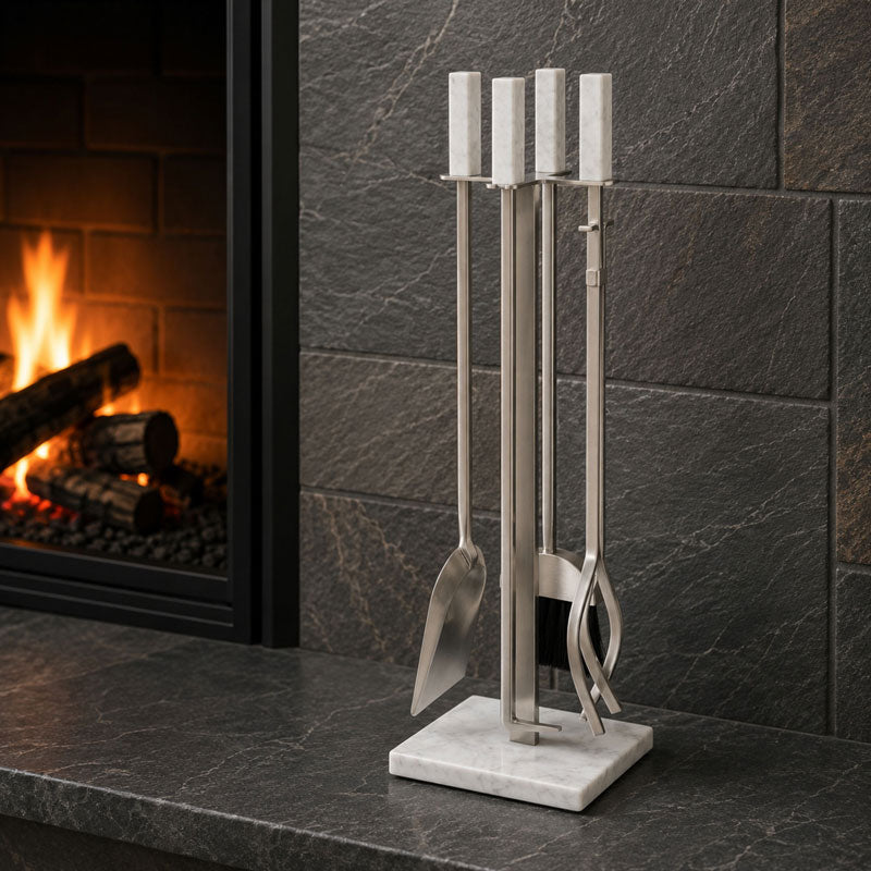 A stainless steel fireplace tool set with white marble handles and a white marble base, displayed on a dark stone fireplace mantel next to a burning fire.