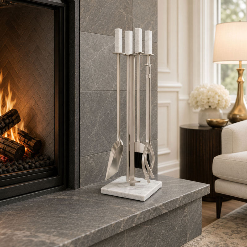 A stainless steel fireplace tool set with white marble handles and a matching marble base, displayed on a gray stone hearth in front of a lit fireplace in a modern living room setting.