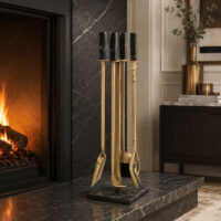 A set of fireplace tools including a poker, shovel, and chisel, finished in polished gold with black accents, displayed on a dark stone mantel in front of a lit fireplace.