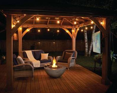 Outdoor wooden patio cabana at night with warm string lights, a fire pit table, wicker seating, and a large outdoor movie screen in the background.