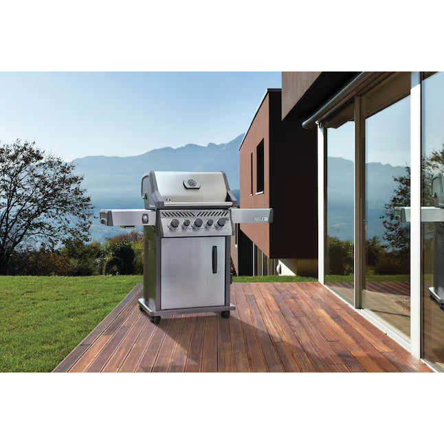 A stainless steel gas grill with side burners is shown on a wooden deck next to a modern house, with a grassy lawn and mountains in the background.