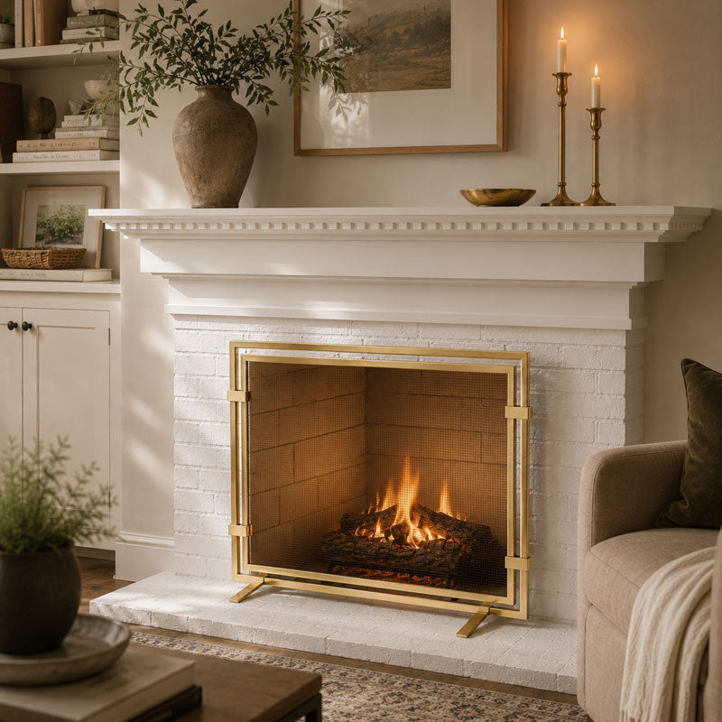 A gold fireplace screen with legs stands in front of a white brick fireplace with a lit fire, set in a cozy living room with a mantel, candles, and a sofa.
