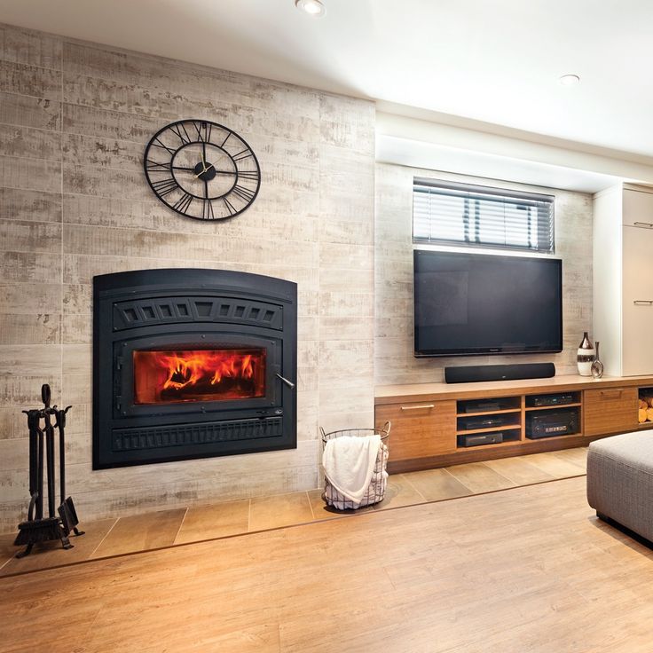 A black wood-burning fireplace with a glowing fire, set into a light stone wall in a modern living room. A large black Roman numerus wall clock hangs above the fireplace, and a wooden media console with a television and soundbar is to the right.