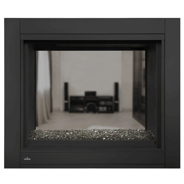 A modern black electric fireplace insert with a reflective glass window showing a blurred interior room, a bed, and a nightstand, with decorative gray and silver pebbles at the base. A small 'Simpson' brand logo is visible in the bottom-left corner.