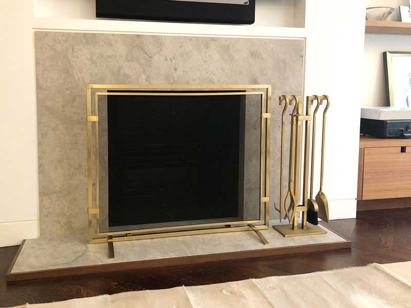 Gold metal fireplace screen and matching fireplace tools set on a stone fireplace hearth in a home interior.